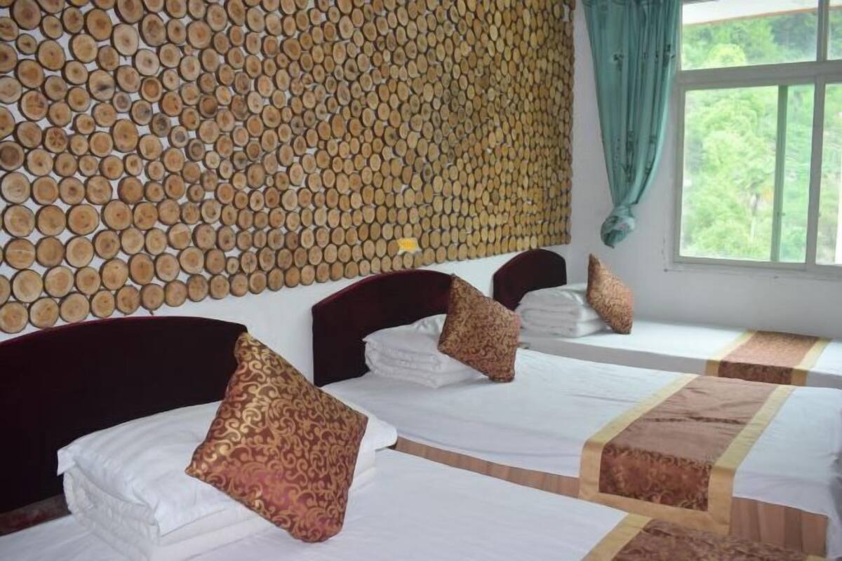 A hotel room with three beds, each with white linens and patterned pillows. The room features a wooden wall panel design and a large window with a view of greenery.