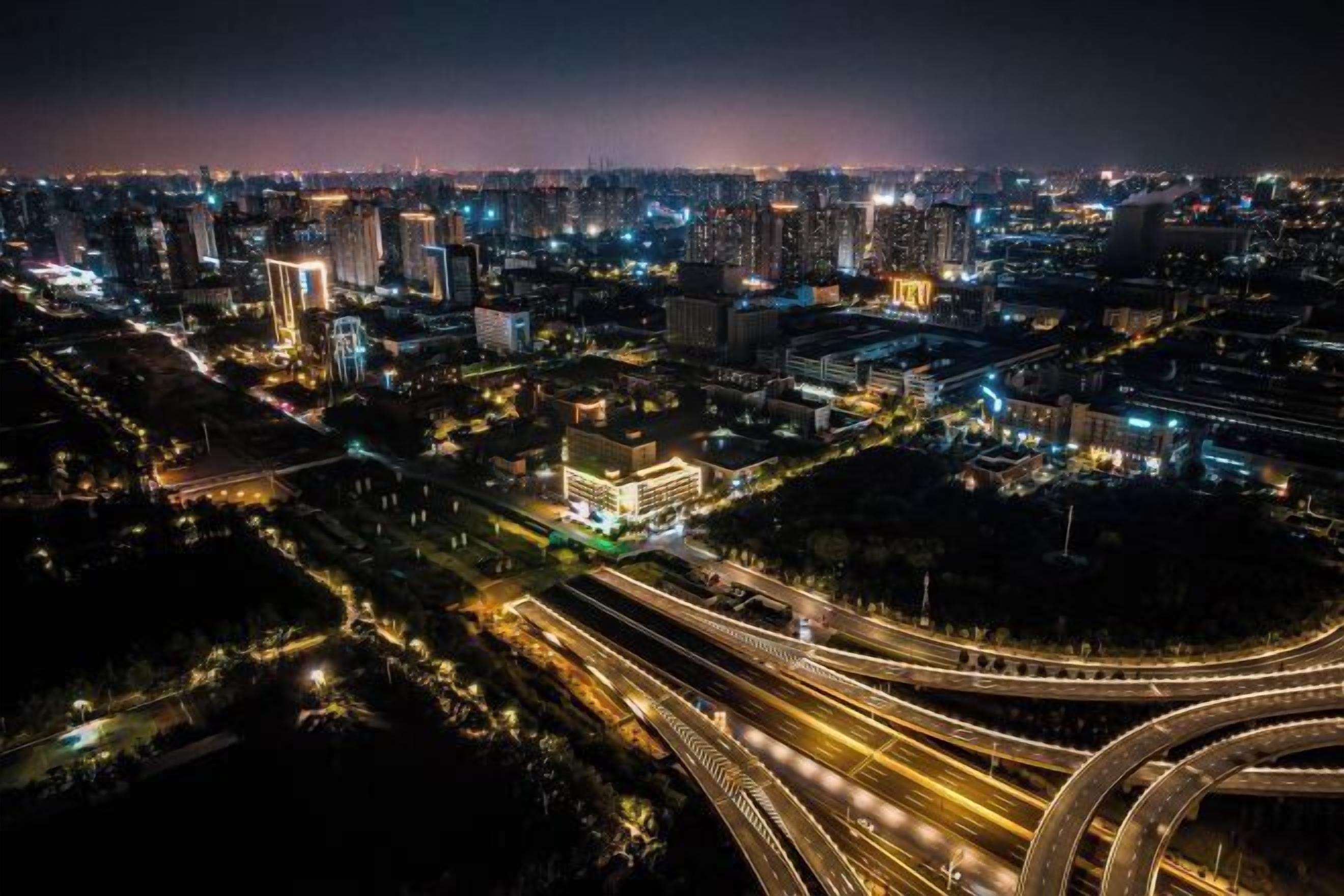 A nighttime aerial view of a cityscape with multiple high-rise buildings, interconnected roads, and a prominent roundabout.