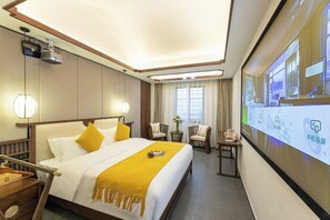 Una habitación de hotel con cama, mesitas de noche, un escritorio y una pantalla grande mostrando contenido.