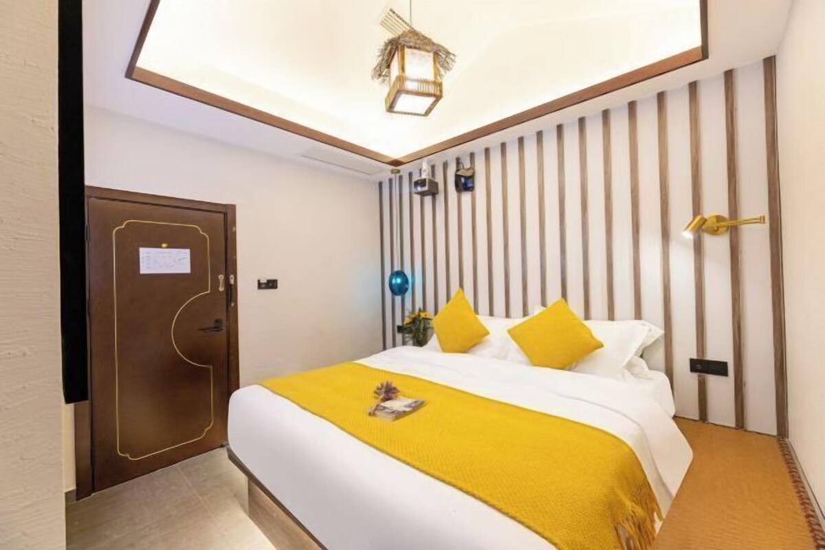 Una habitación de hotel con una cama, una puerta de madera, una lámpara fijada a la pared y una luz en el techo.
