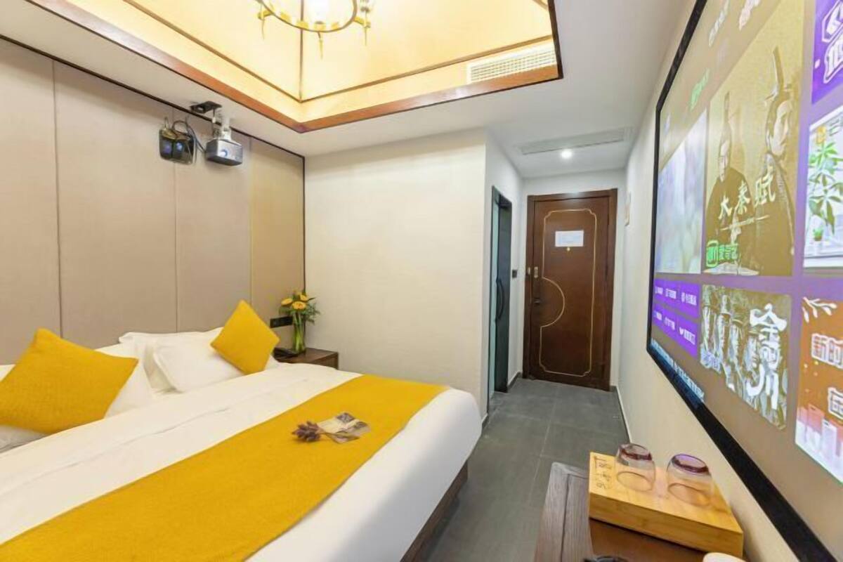 Habitación de hotel con cama, mesita de noche, proyector montado en la pared y puerta que da a otra habitación.