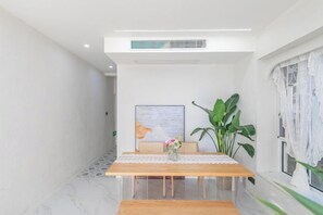 A dining area with a wooden table, white walls, and a large window with white curtains.