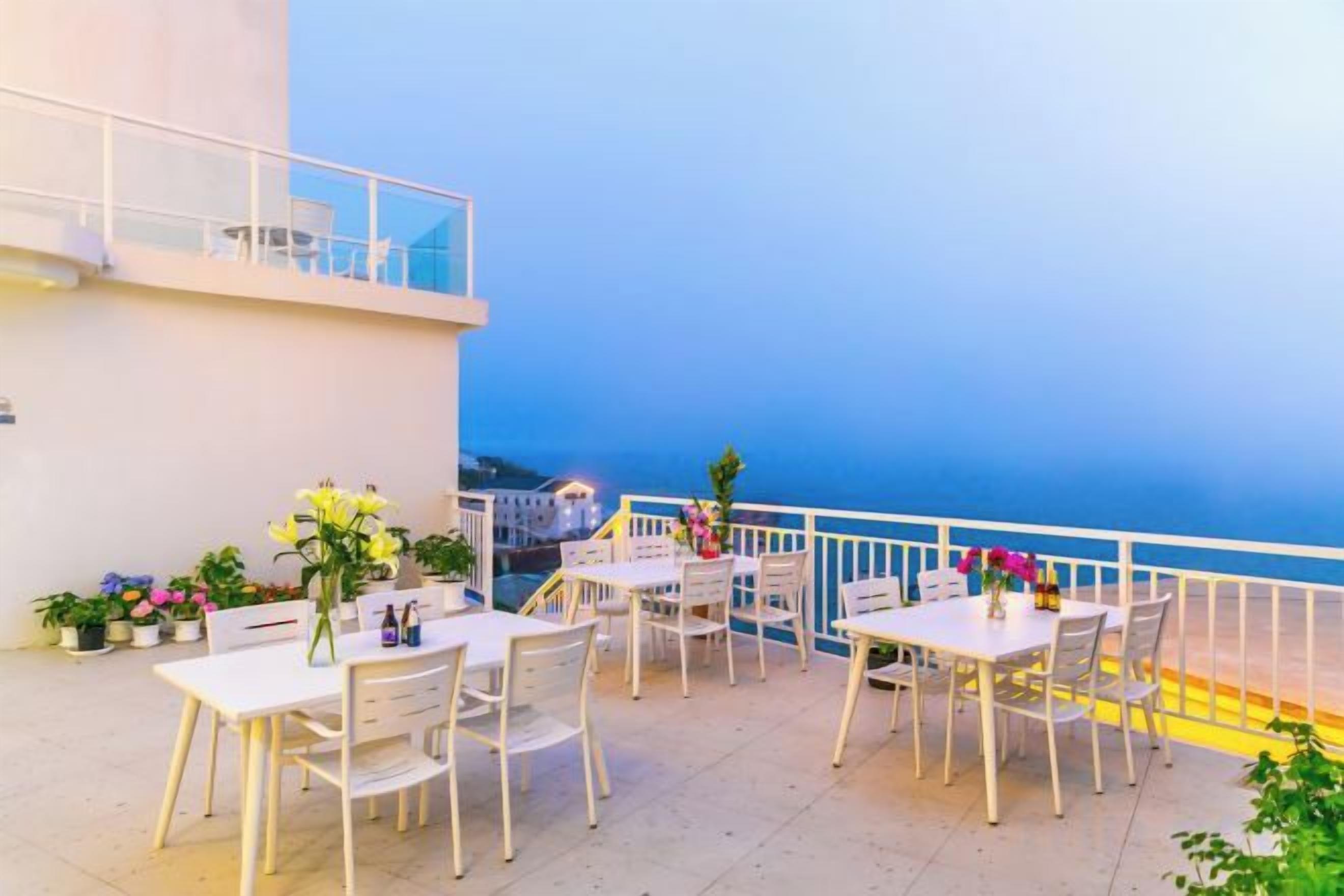 An outdoor terrace with white tables and chairs, potted plants, and a view of the sea.