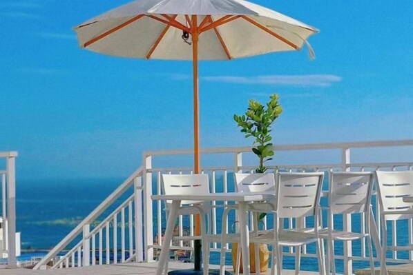 A deck with white chairs and a table, an umbrella, and a railing overlooking the ocean.