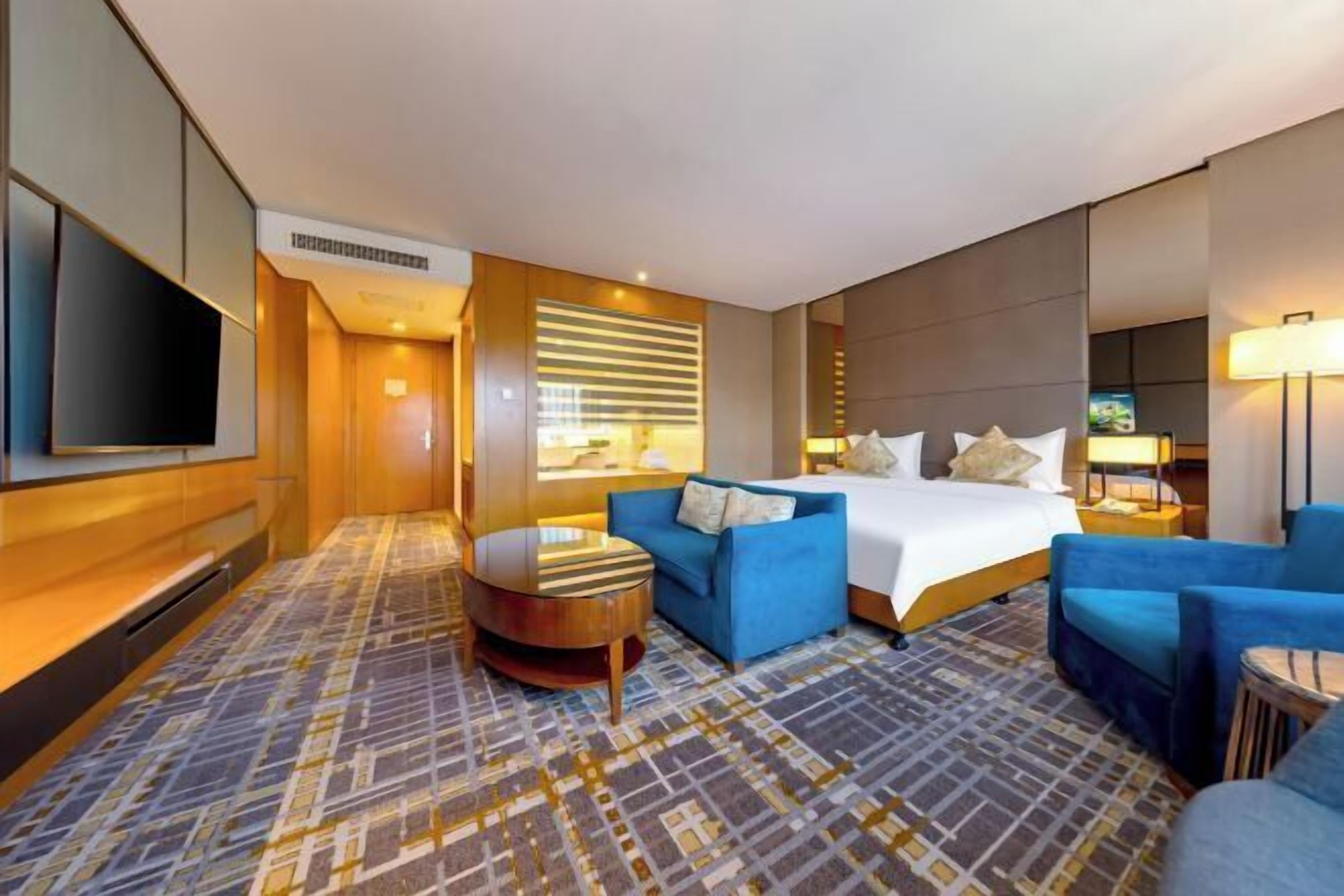 Habitación de hotel con cama, sofá azul, una pequeña mesa redonda y televisor montado en la pared.