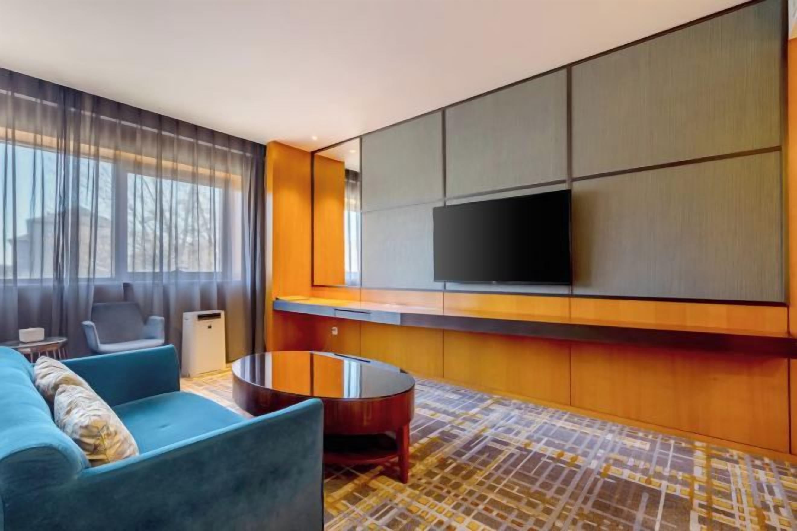 Habitación de hotel moderna con un televisor de pantalla plana grande montado en la pared, un sofá azul, una mesa redonda de centro y una alfombra estampada.