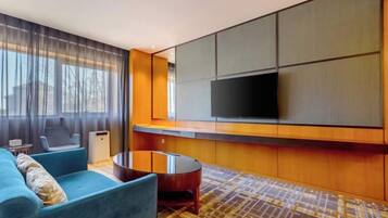 Habitación de hotel moderna con un televisor de pantalla plana grande montado en la pared, un sofá azul, una mesa redonda de centro y una alfombra estampada.