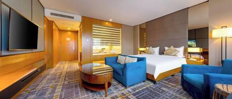 Een hotelkamer met een bed, twee blauwe fauteuils, een kleine ronde tafel, een televisie en een kamerbreed tapijt met een patroon.