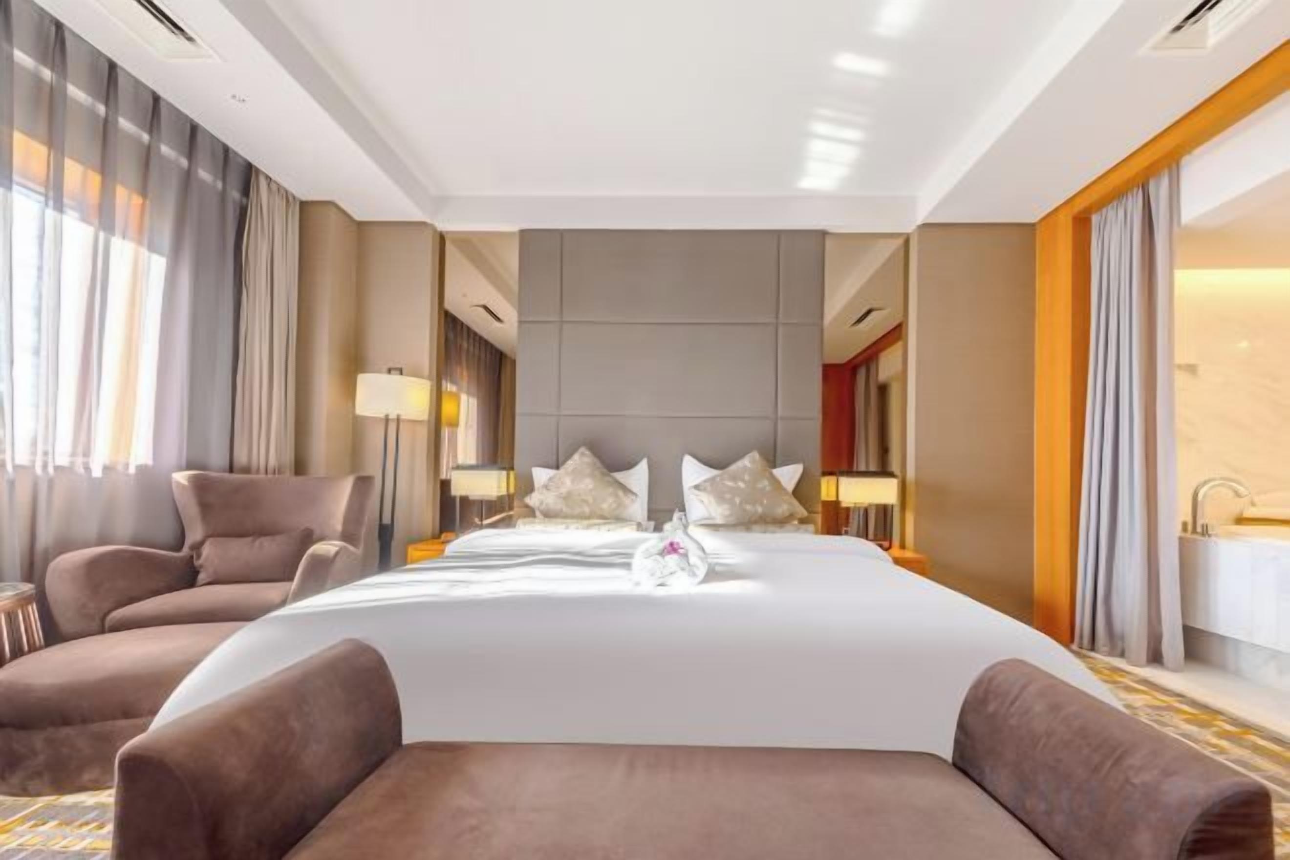Habitación de hotel con una cama grande, dos mesitas de noche, una zona de estar con una silla y un baño con lavabo y espejo.