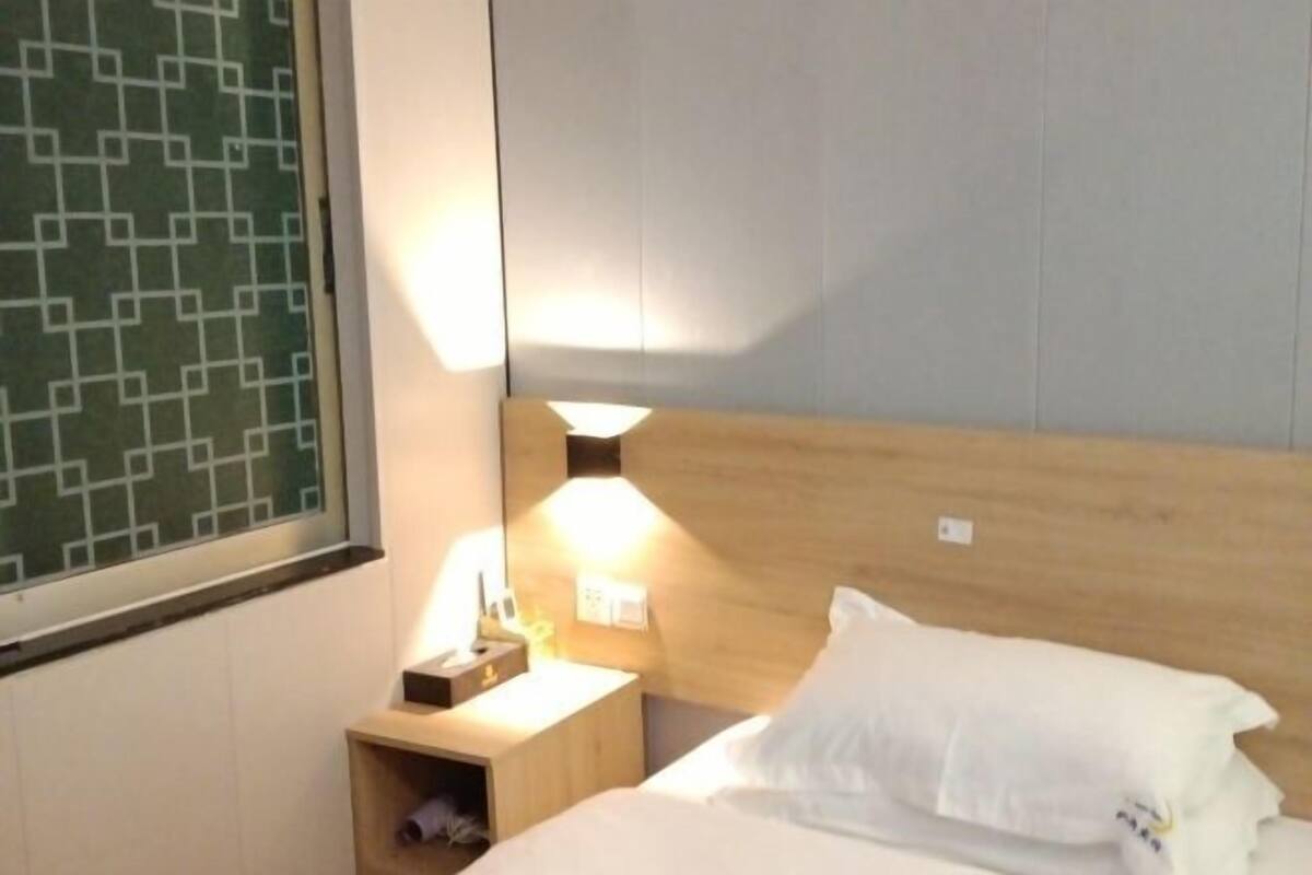 A hotel room with a wooden headboard, a bedside table with a lamp, and a window with a geometric pattern.
