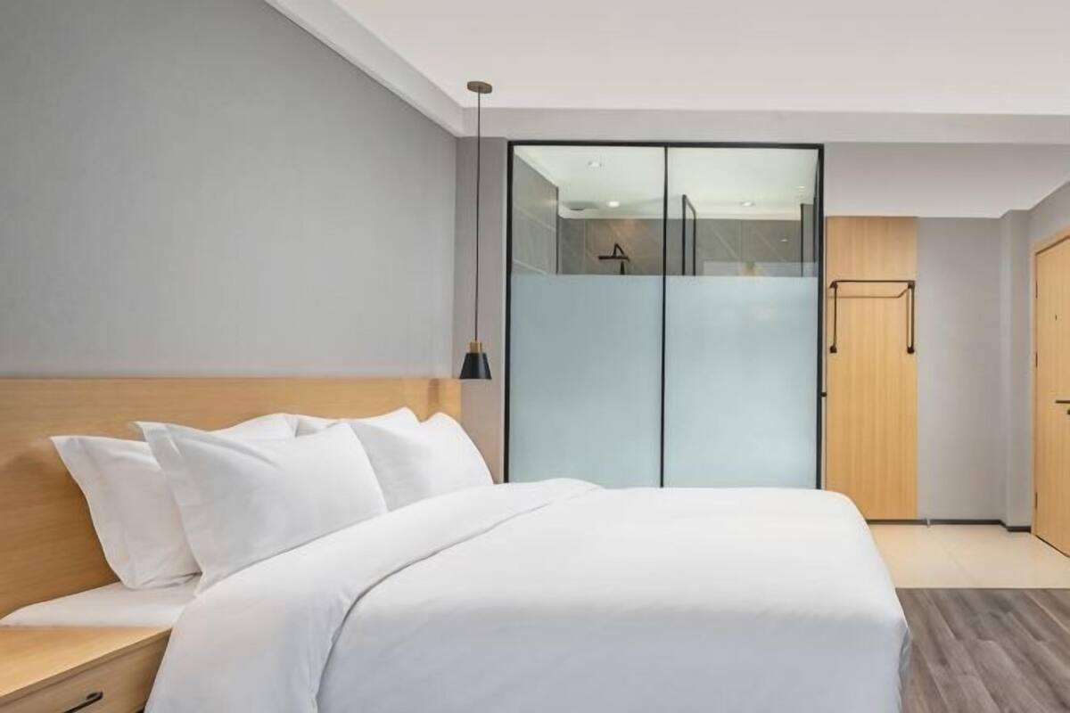 A modern hotel room with a large bed, a wooden nightstand, a wardrobe with a mirror, and a hanging light fixture.