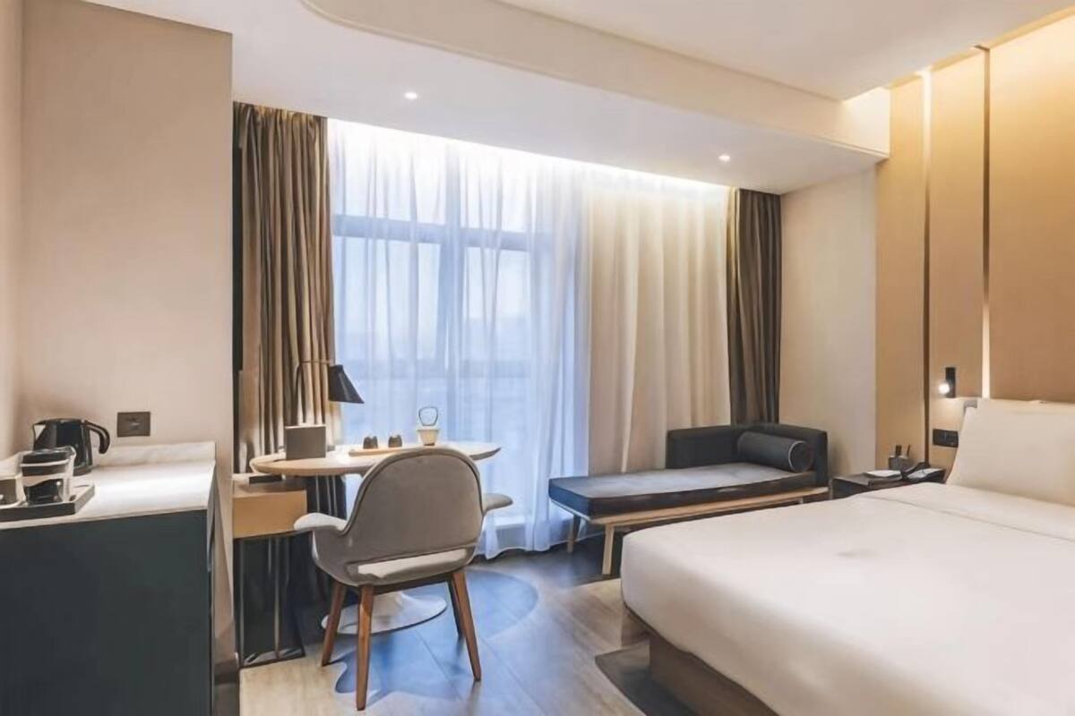 A hotel room with a bed, a desk with a chair, a small table, and a window with curtains.