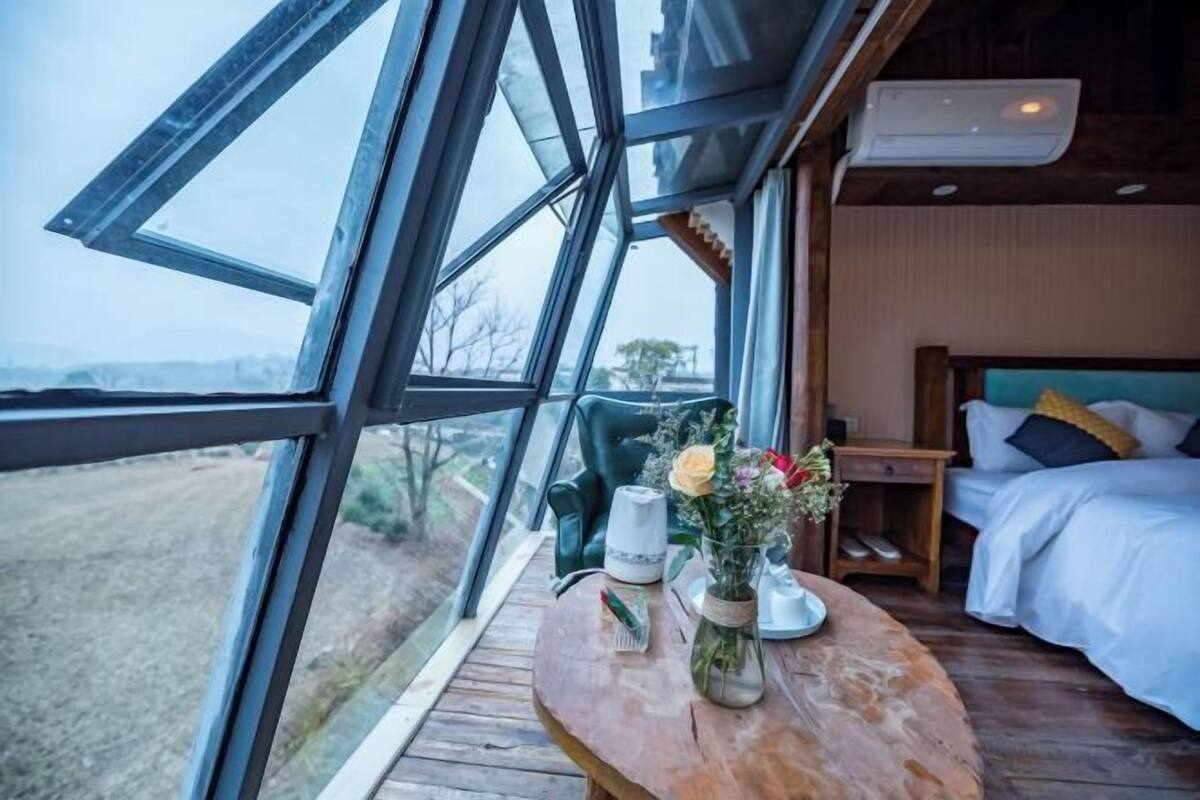 A room with a large window, a wooden table with a vase of flowers, a bed with pillows, and a nightstand.