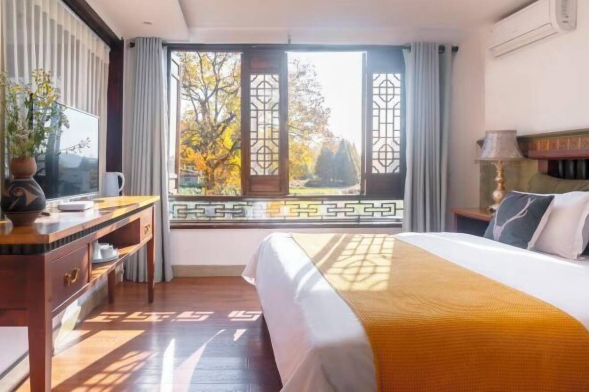 A hotel room with a bed, a desk, a chair, a window with a view of trees, and a lamp.