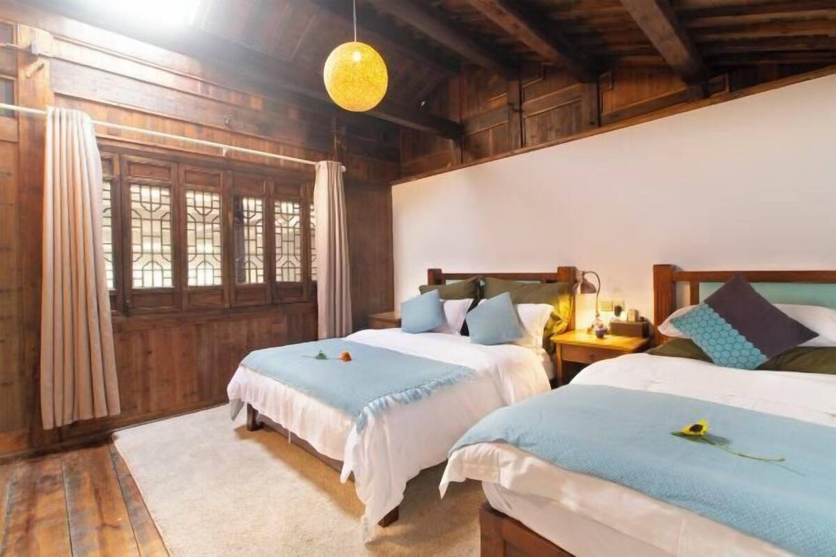 A room with two beds, wooden walls, a hanging light fixture, and a window with a curtain.
