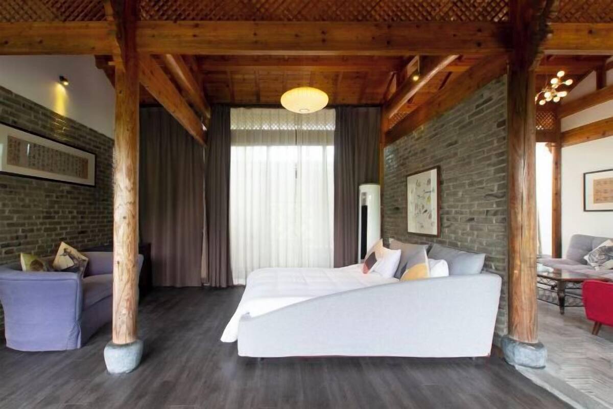 A room with a bed, wooden beams, a stone wall, a window with curtains, and a sofa.