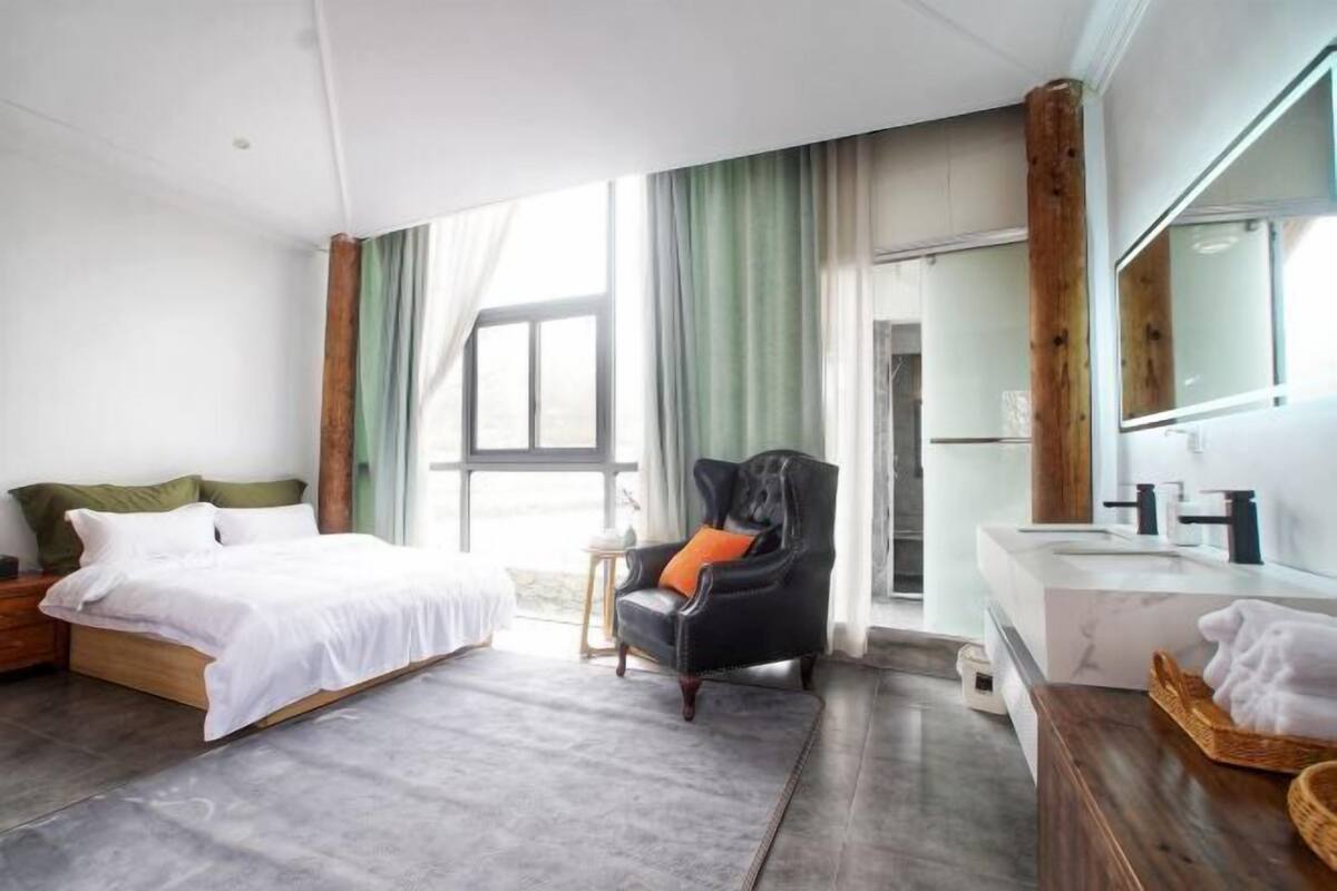 A modern hotel room with a bed, a chair, a vanity with a mirror, and a large window.