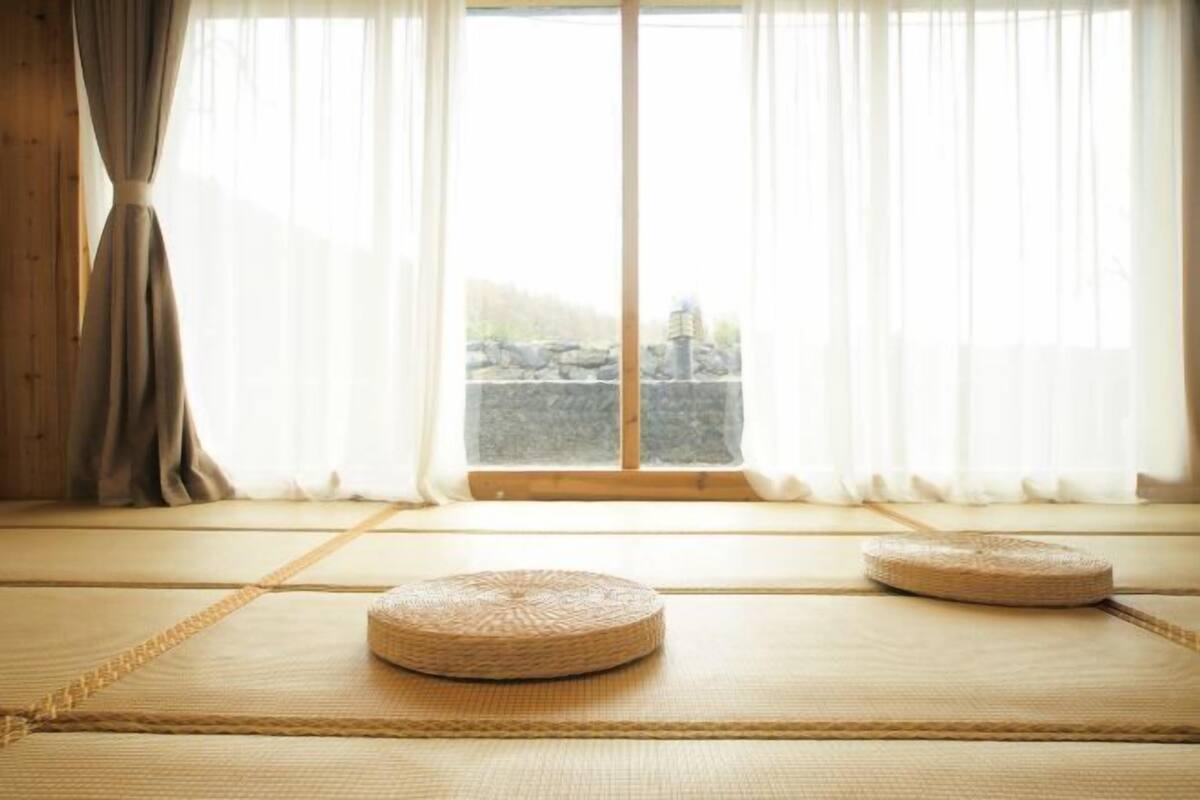 A tatami mat room with a view of a stone wall and a window with sheer curtains.