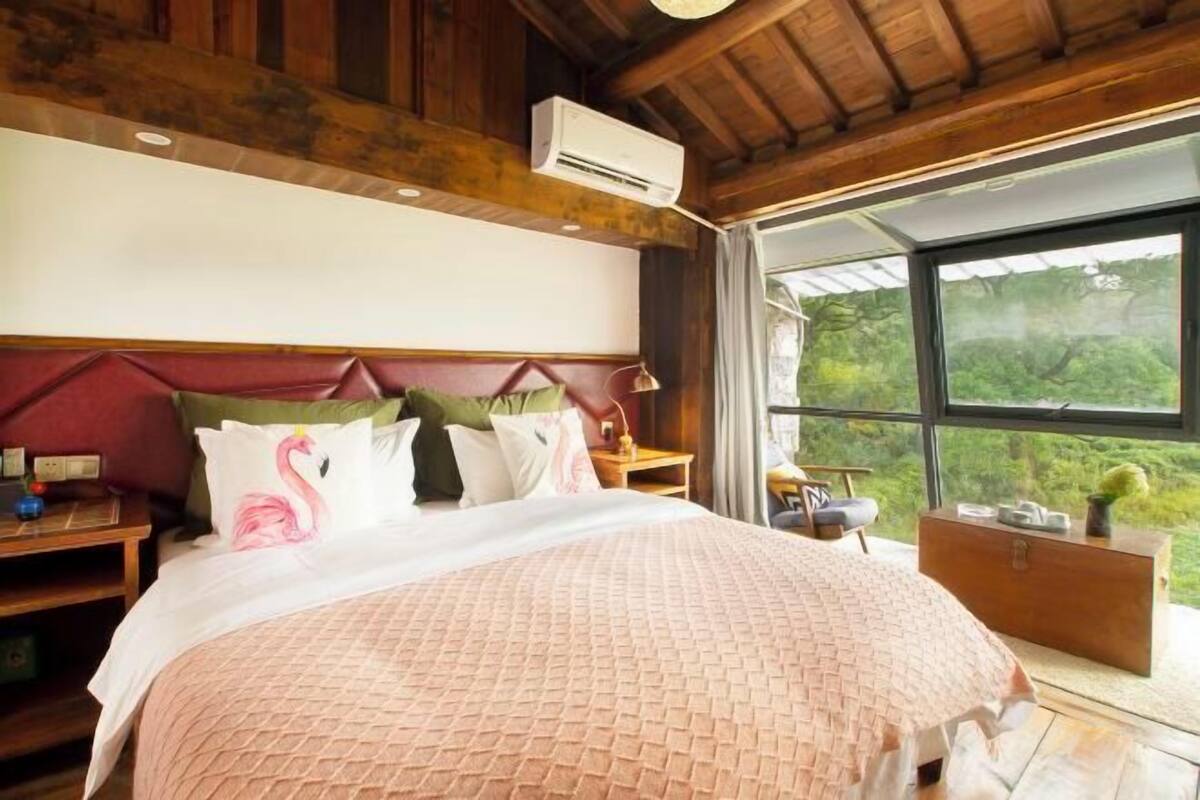 A bedroom with a large bed, a wooden ceiling, an air conditioning unit, a nightstand, a dresser, and a window with a view of greenery.