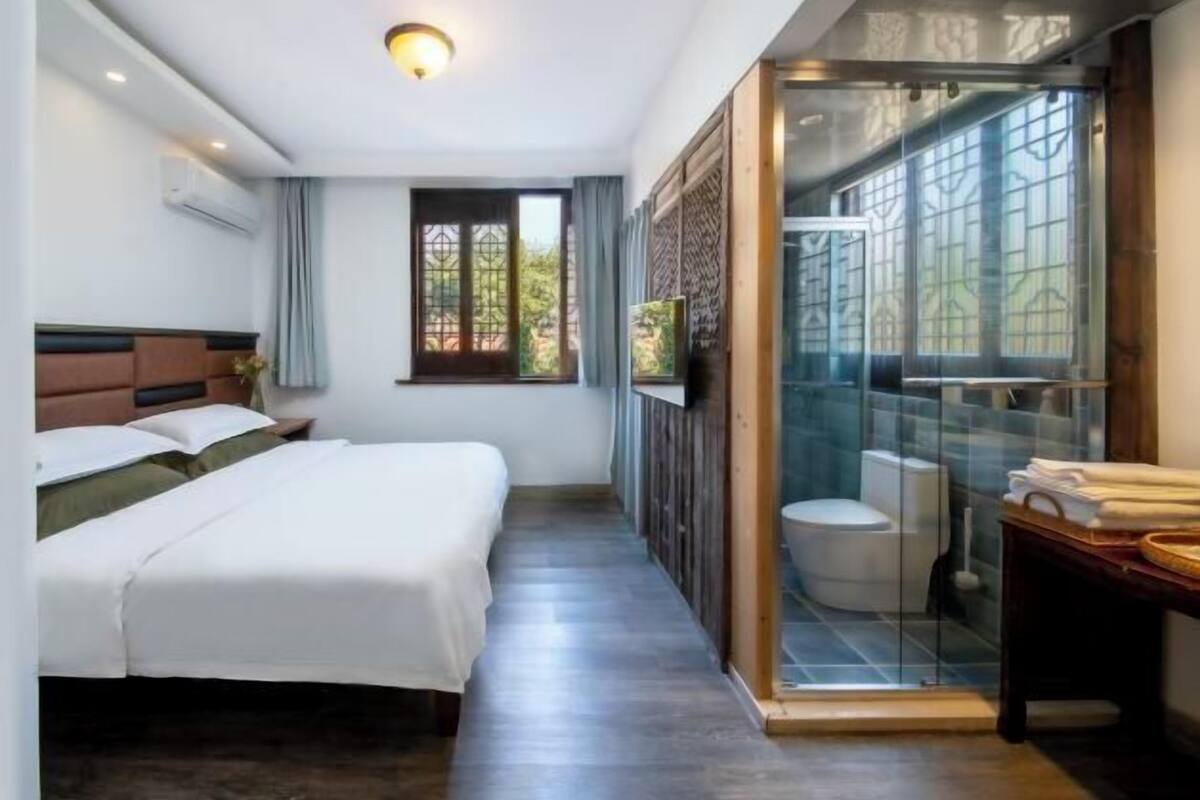 A hotel room with a bed, a shower area with glass doors, and a window with curtains.