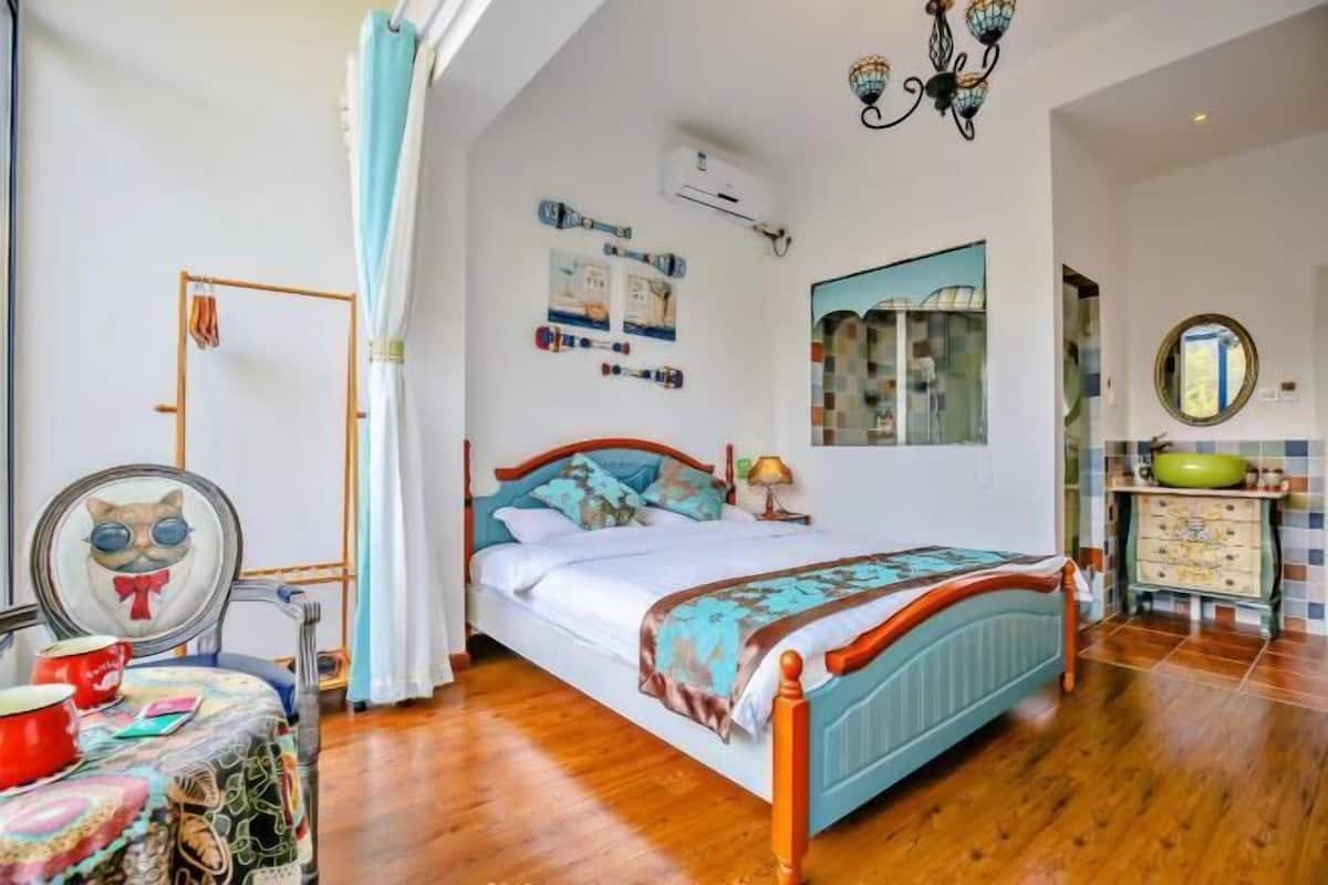 Ein Schlafzimmer mit einem hölzernen Bett, einem Nachttisch, einem Spiegel, einem Fenster mit Vorhängen sowie einem Badezimmer mit Waschbecken und Spiegel.