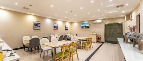A hotel dining area with multiple tables and chairs, a buffet counter, and a television screen on the wall.