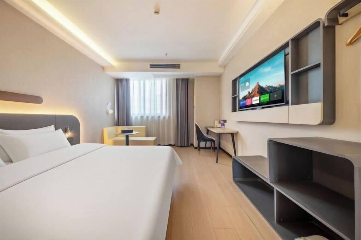 Ein Hotelzimmer mit Bett, Schreibtisch, Stuhl, einem an der Wand befestigten Fernseher und einem Fenster mit Vorhängen.