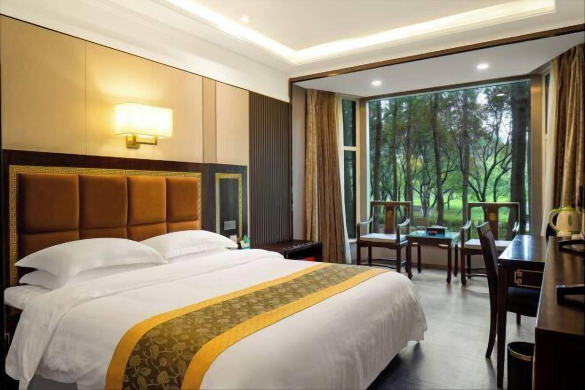 A hotel room with a large bed, a desk, and a view of trees through the window.