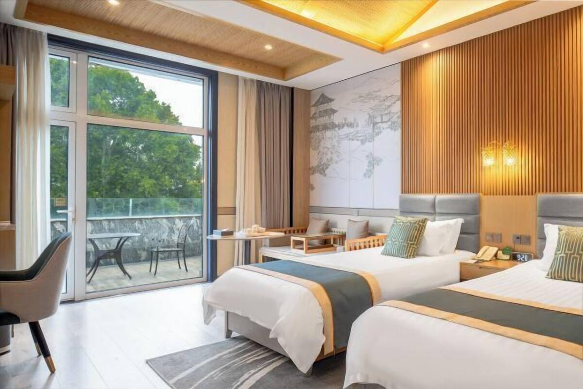 A modern hotel room with two beds, a desk, and a balcony with a view of trees.
