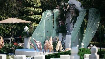 An outdoor event stage with a backdrop of greenery and flowers, surrounded by white chairs and tables.