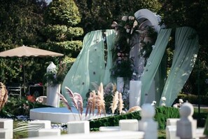 An outdoor event stage with a backdrop of greenery and flowers, surrounded by white chairs and tables. - Yujing Garden Holiday Hotel (Shijiazhuang)