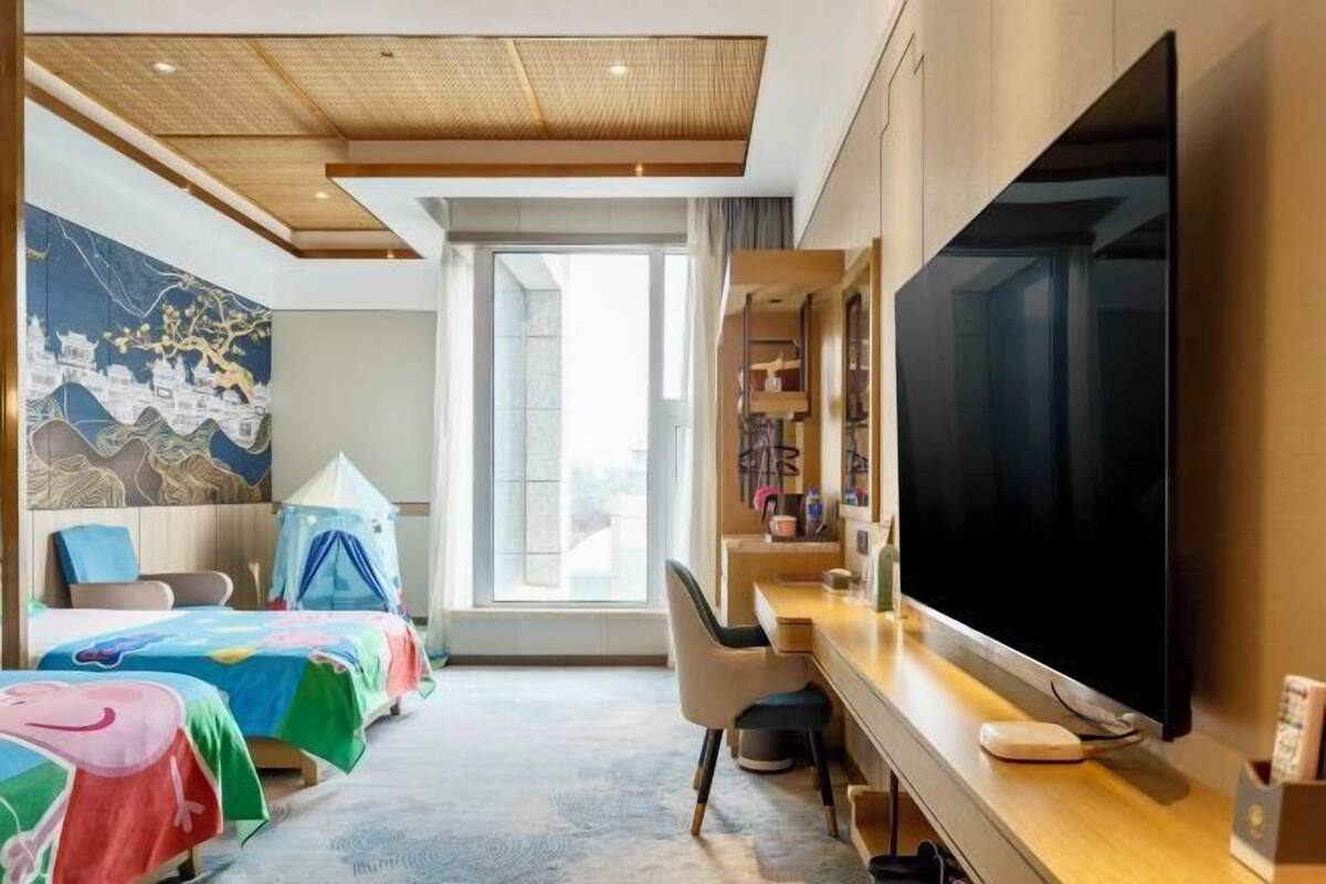 A hotel room with a large bed, a desk with a flat-screen TV, a chair, and a large window.