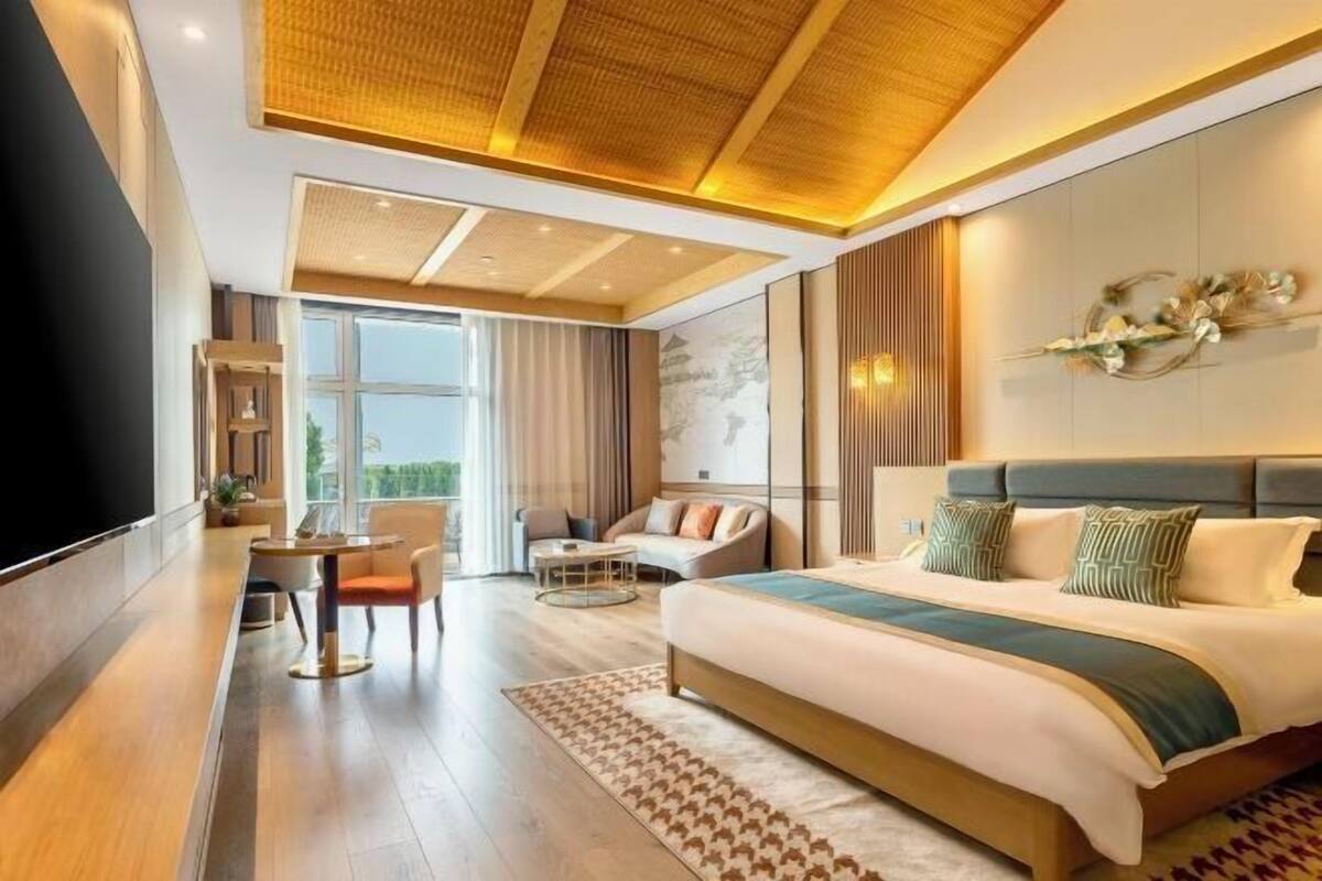 A modern hotel room with a large bed, a seating area with a sofa and chairs, a dining table, and a large window providing a view.