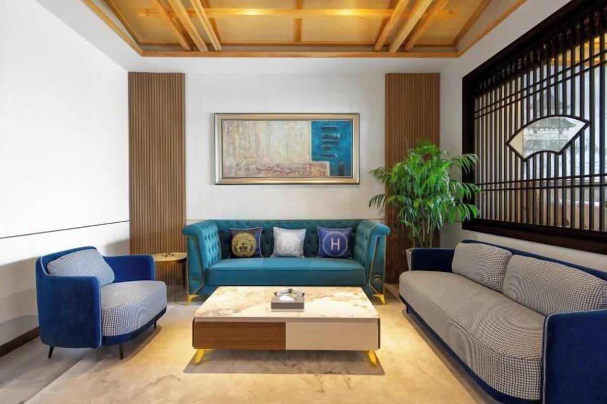 A modern living room with a teal sofa, a blue armchair, and a patterned sofa. There is a coffee table in the center, a painting on the wall, and a potted plant.