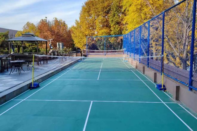 An outdoor tennis court with a net, surrounded by a blue fence and trees with autumn foliage.