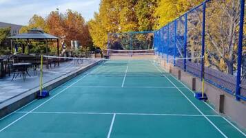 An outdoor tennis court with a net, surrounded by a blue fence and trees with autumn foliage.