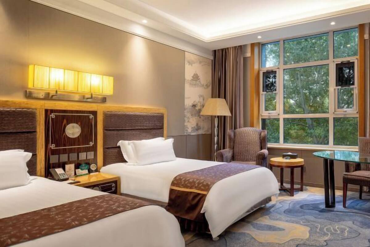 A hotel room with two beds, a sitting area, a small table, and a large window with a view of trees.