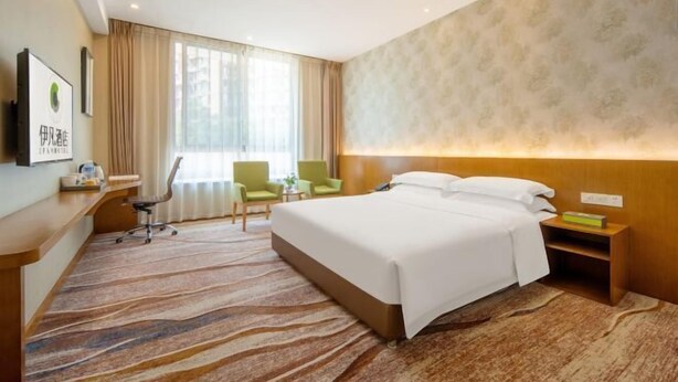 Ein Hotelzimmer mit einem großen Bett, einem Schreibtisch mit Stuhl, einem kleinen Tisch und einem Fenster mit Vorhängen.