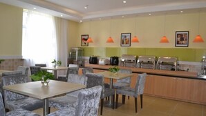 Un comedor de hotel moderno con sillas grises acolchadas, una larga barra con electrodomésticos de cocina y lámparas colgantes naranjas.