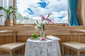 A room with wooden walls, a round table with a floral tablecloth, two chairs, a potted plant, a vase with flowers, and a view of traditional architecture through a window.