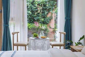 A hotel room with a bed, two wooden chairs, a round table with a vase of flowers, and a view of greenery outside.