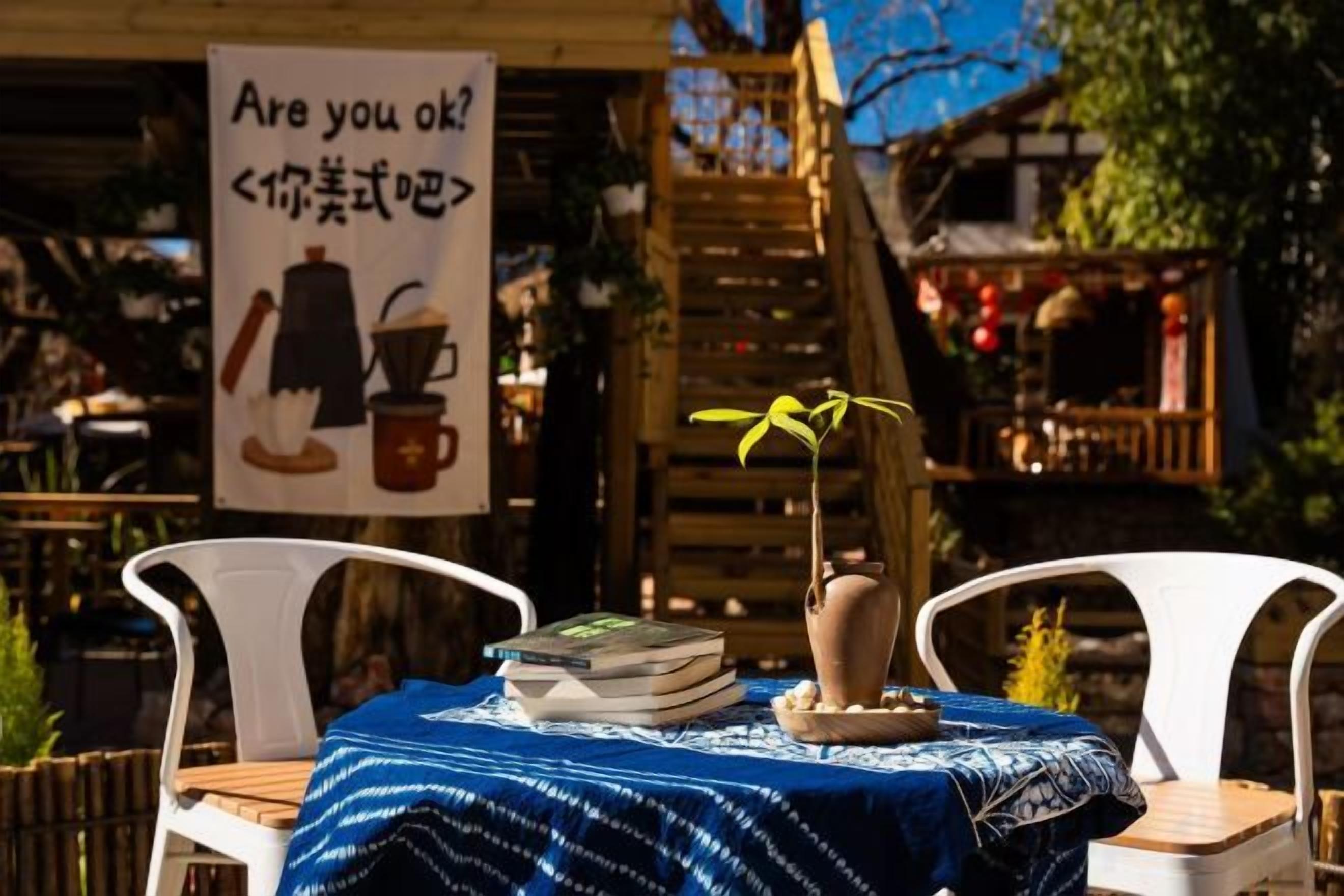 An outdoor seating area with a table covered by a blue cloth, a sign with a coffee cup illustration, and a wooden staircase in the background.
