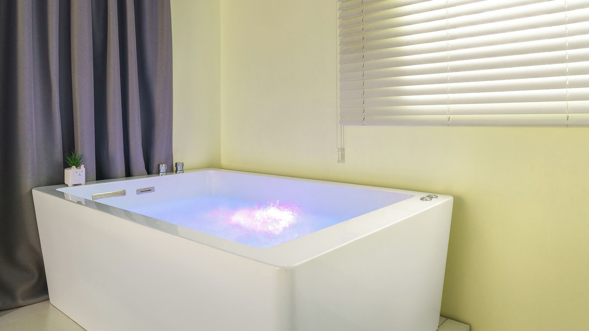 Une salle de bain moderne avec une baignoire blanche, une plante en pot et une fenêtre avec des stores.