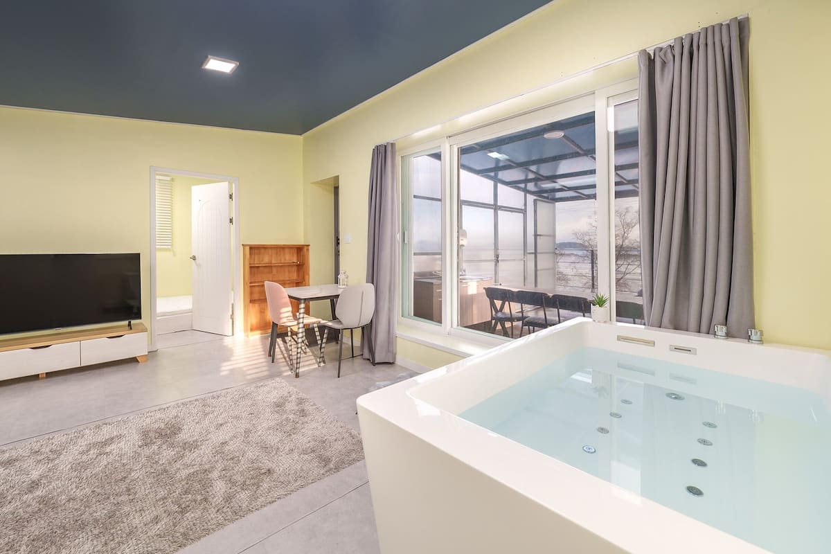 A modern living room with a flat-screen TV, a dining area with a table and chairs, a large window with curtains, and a jacuzzi tub.