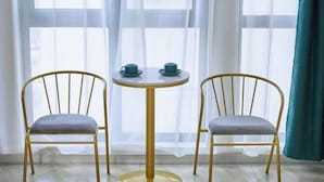 Een kamer met twee stoelen en een kleine ronde tafel, een raam met transparante gordijnen en een blauwgroene gordijn aan de rechterkant.