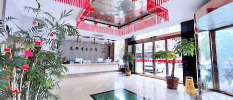 A hotel lobby with a reception desk, potted plants, and a large window.