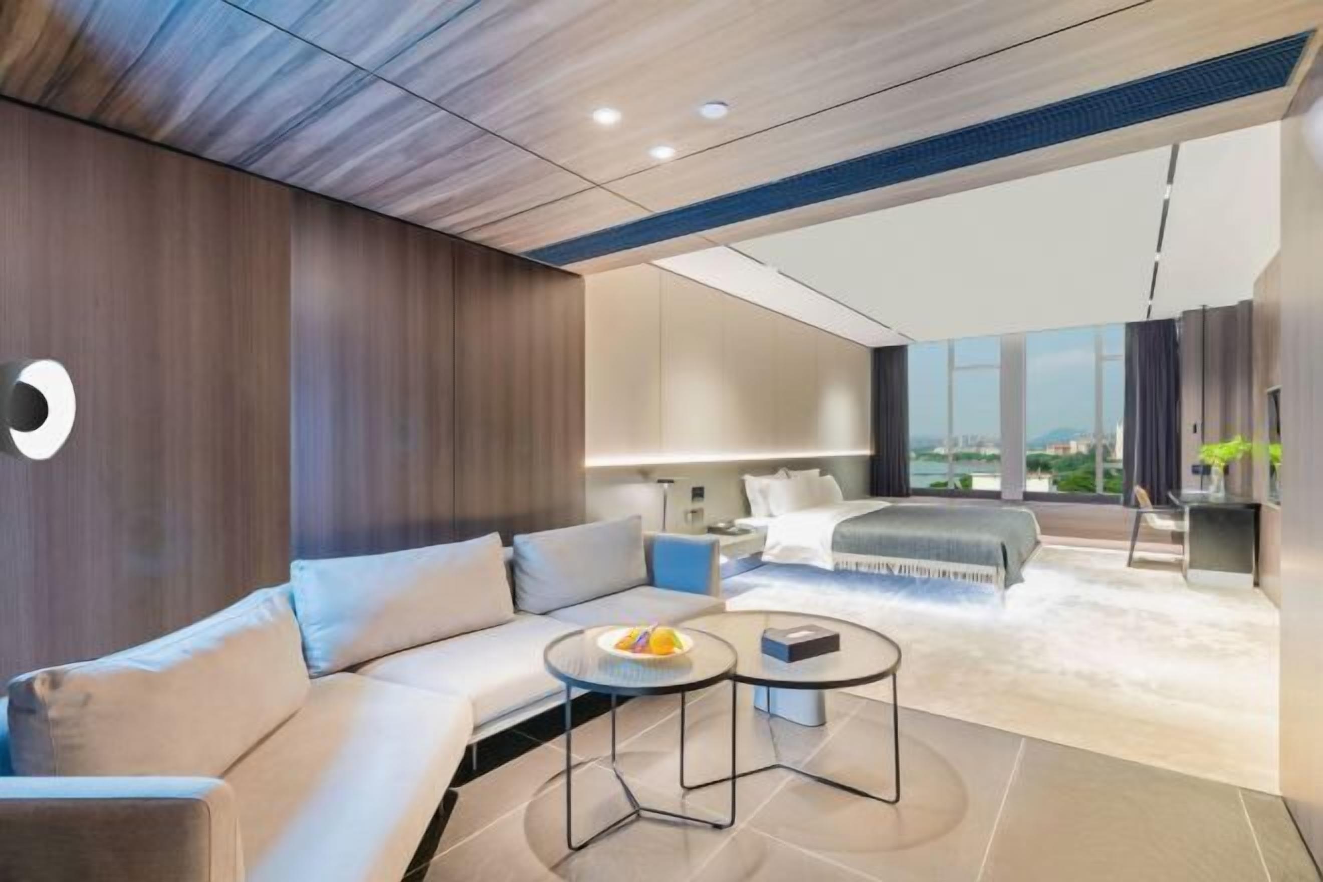 A modern hotel room with a sofa, a round coffee table, a bed, and a view of the city.
