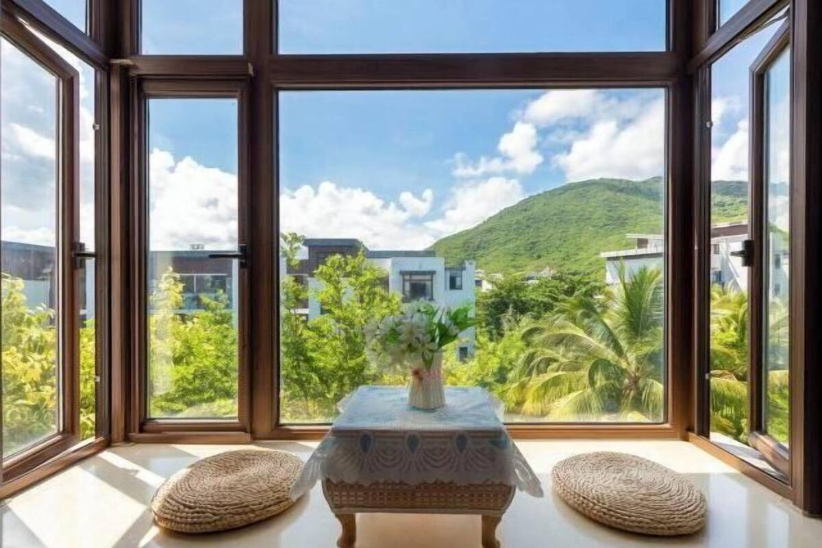 Une pièce avec une grande fenêtre, une petite table sur laquelle est posé un vase, et deux coussins.