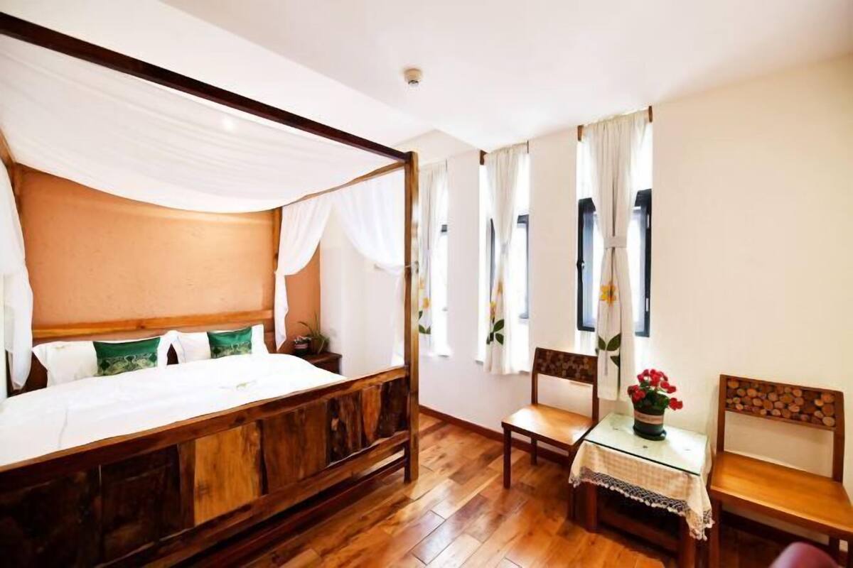 A room with a four-poster bed, a wooden table, a chair, and a small table with a vase of flowers.