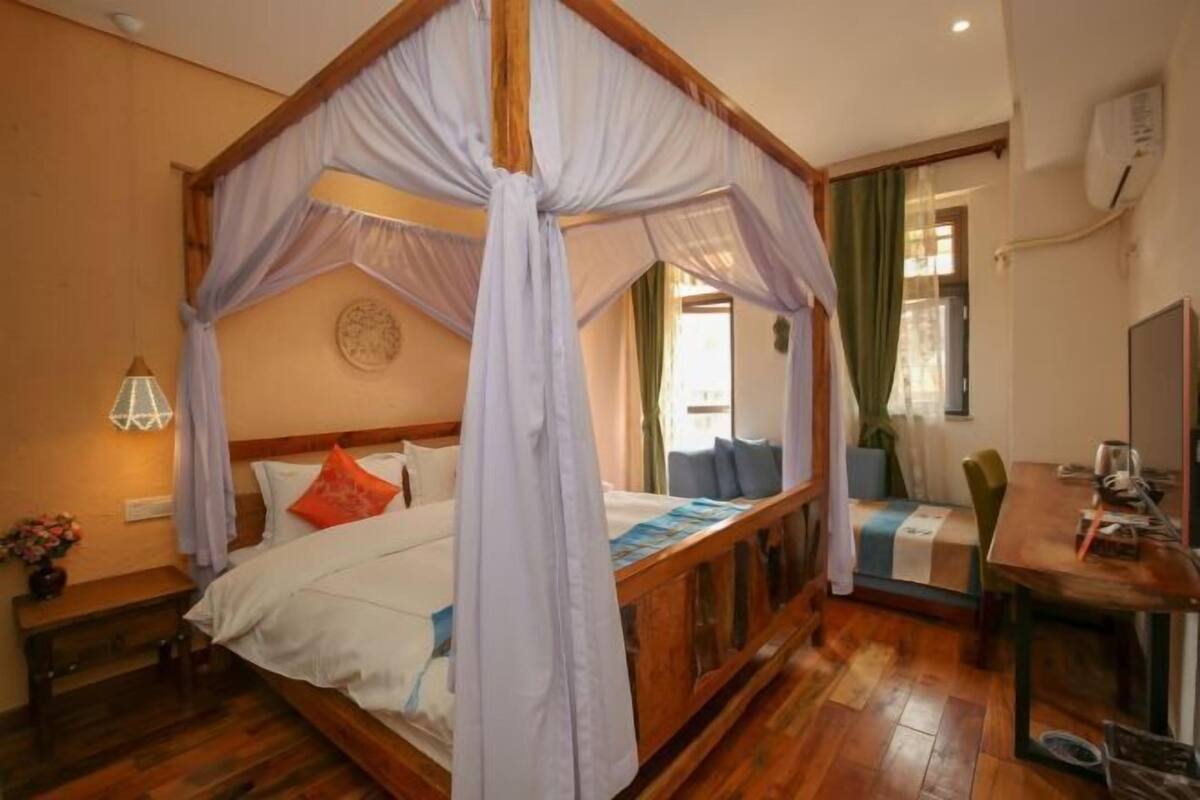 A four-poster canopy bed with a wooden frame, a bedside table with a lamp, and a view of the room's interior.